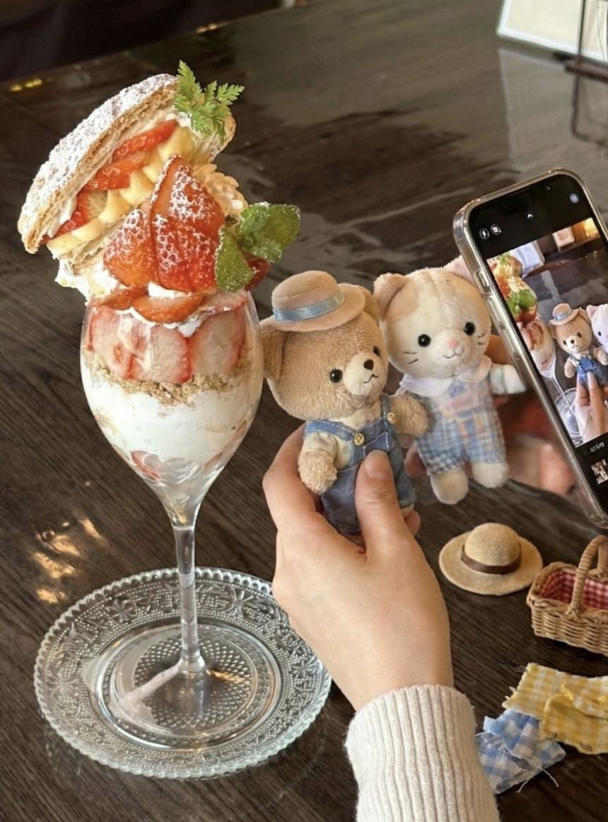 テーブルに並べたぬいぐるみと、いちごのパフェ。スマートフォンで撮影しているところ